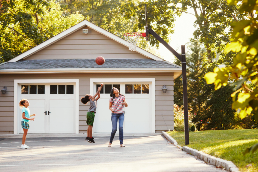 Three people playing basketball in a driveway in front of a residential garage with white double doors.